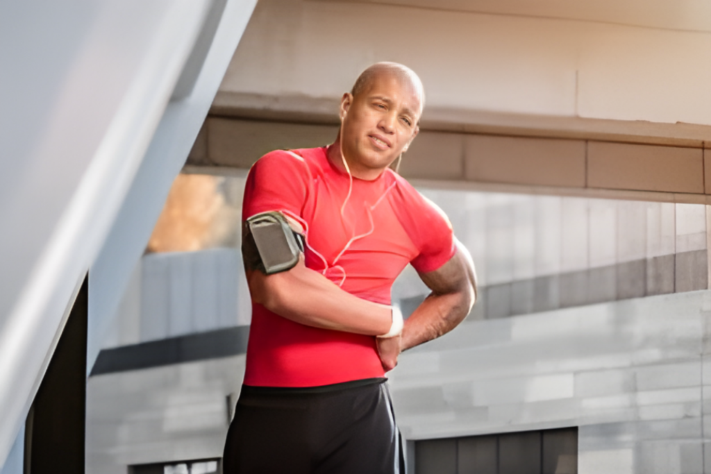 Man in a red athletic shirt stands with hands on hips, wearing a phone armband and earphones outdoors.