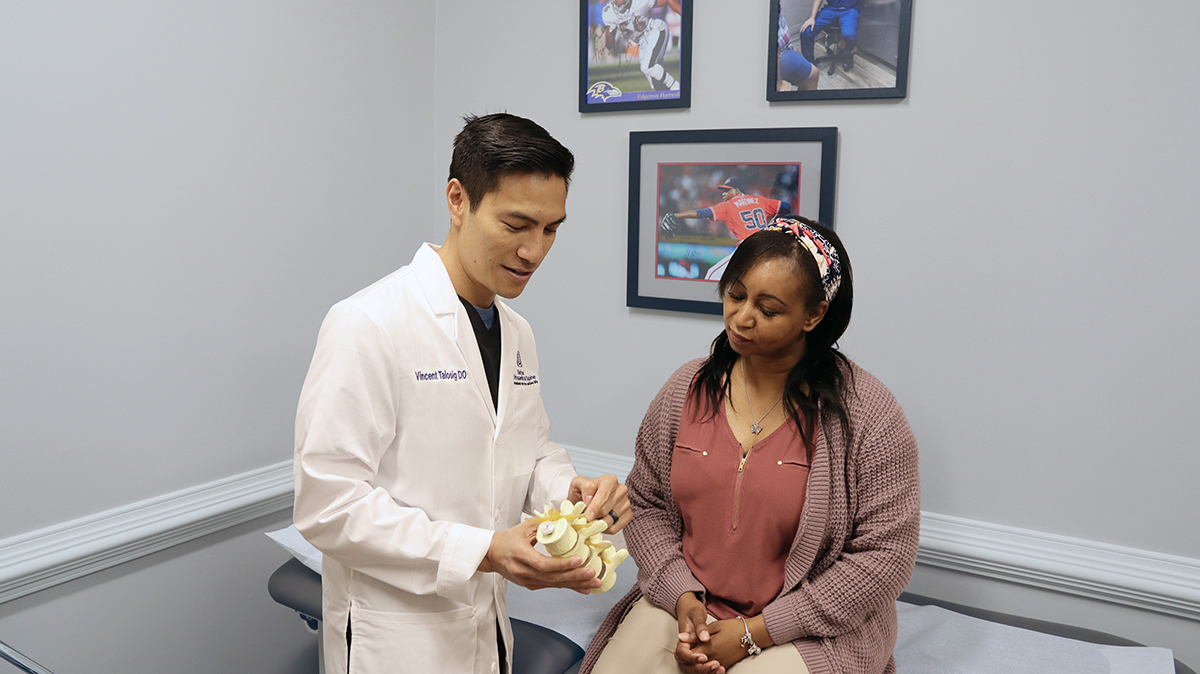 Male doctor in a white coat explains a spine model to a seated female patient in a medical examination room.