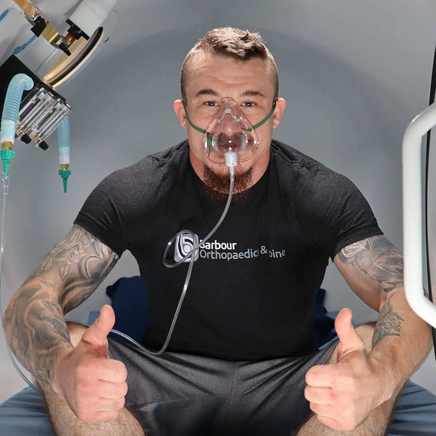 Man with tattoos wearing an oxygen mask inside a clinical chamber, giving thumbs up.