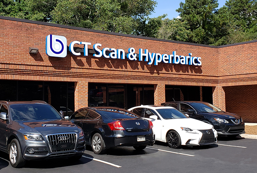 Brick storefront with blue and white sign reading 'CT Scan & Hyperbarics' and several parked cars in front of the building. Details include a red brick exterior and a tree-lined background.