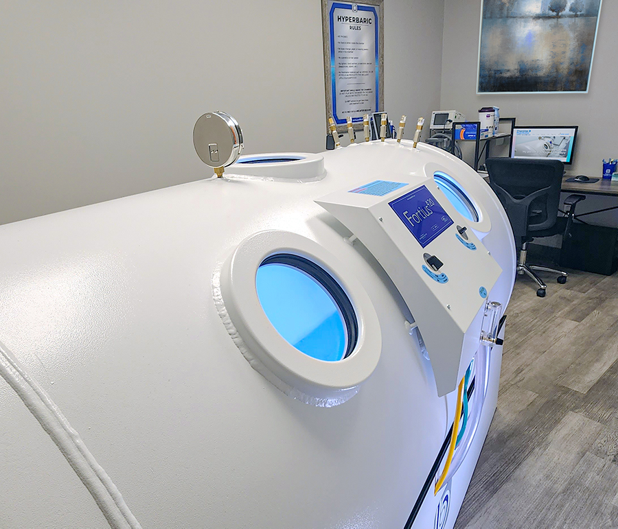 White hyperbaric chamber with blue-lit circular windows and control panel in a clinic treatment room.